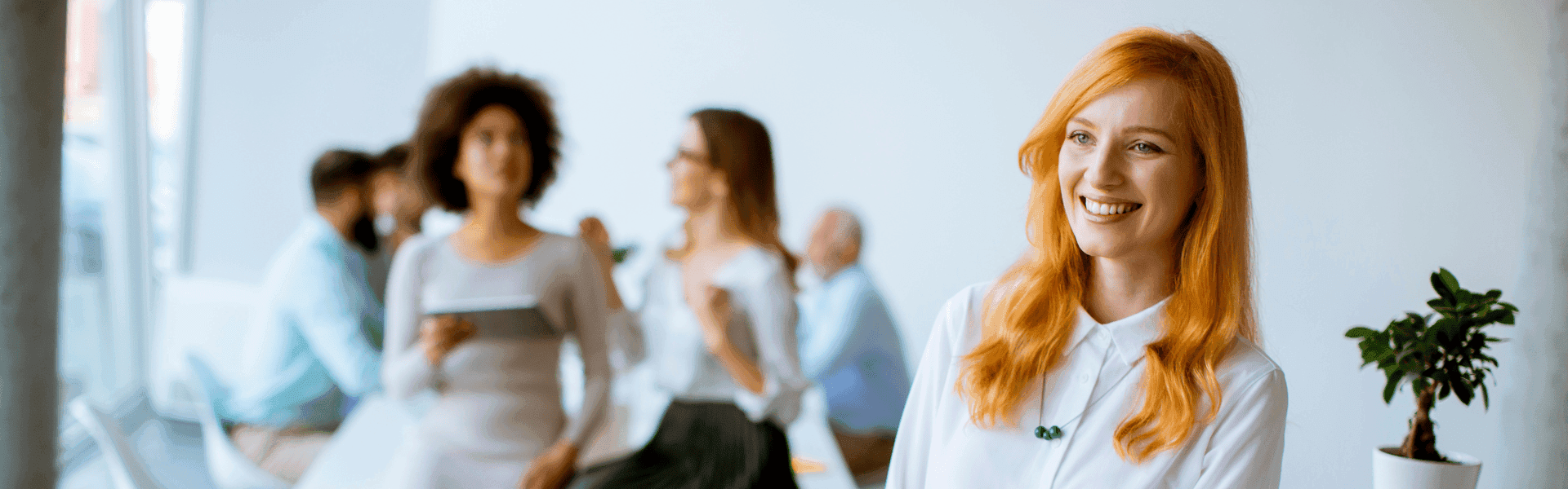 Mitarbeiterin mit langen rötlichen Haaren steht im Büro und lächelt in die Kamera. Im Hintergrund sitzen einige Kolleginnen oder lehnen sich an einen Schreibtisch und unterhalten sich. 