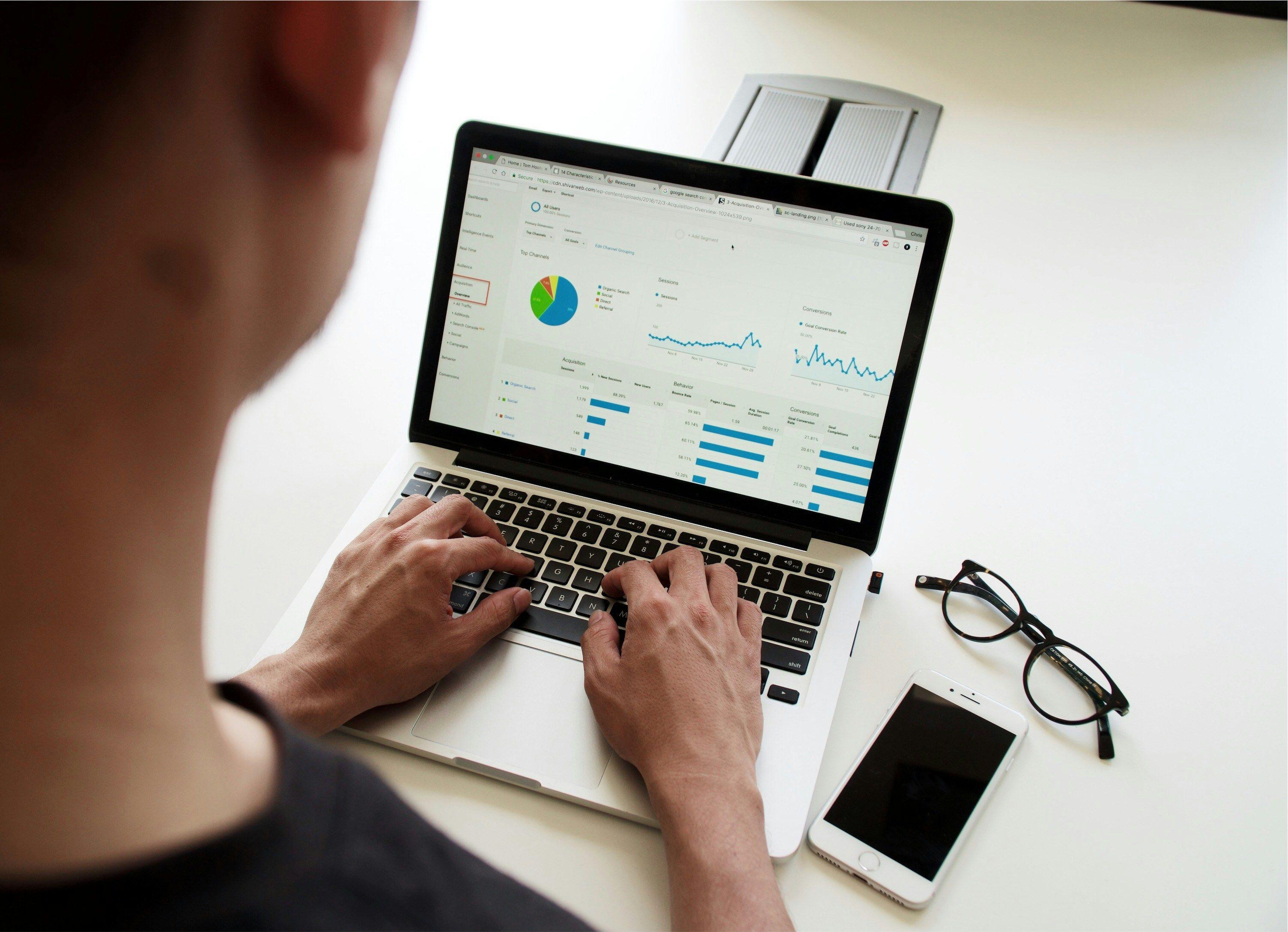 Eine Person arbeitet am Laptop mit Datenanalysediagrammen. Auf dem Tisch liegen eine Brille und ein Smartphone.