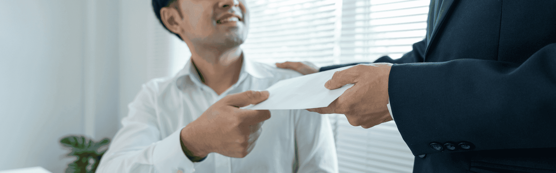 Eine Person im weißen Hemd erhält von einer anderen Person im Anzug einen Briefumschlag überreicht; im Hintergrund sind helle Jalousien und eine Pflanze zu sehen.