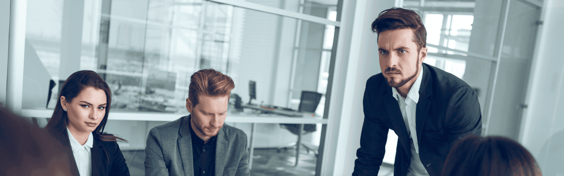 Mehrere Personen in Businesskleidung sitzen in einem modernen Konferenzraum und führen eine Besprechung; eine Person steht und spricht zum Team. Im Hintergrund sind Glaswände und Büros zu sehen.