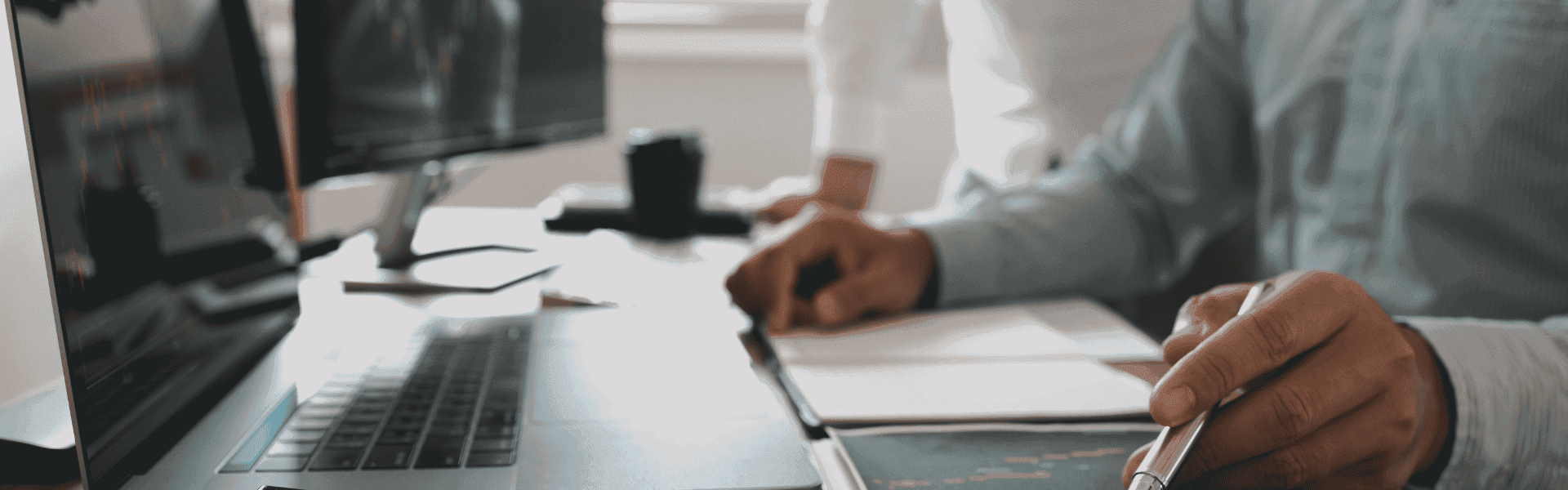 Nahaufnahme von Händen, die auf einem Laptop arbeiten, während jemand im Hintergrund Notizen macht, mit Diagrammen auf dem Bildschirm sichtbar.