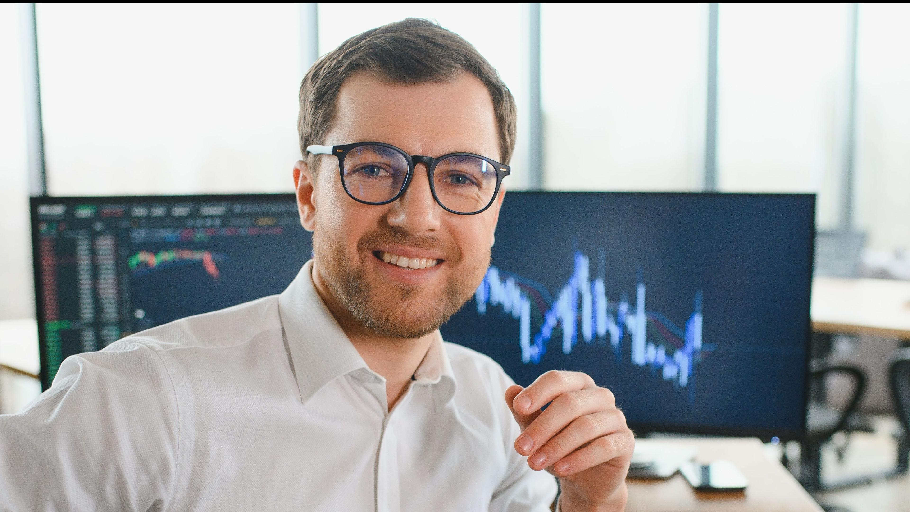 Ein Mann mit Brille lächelt in einem Büro vor einem Monitor mit Kursdiagrammen. Bürofenster im Hintergrund.