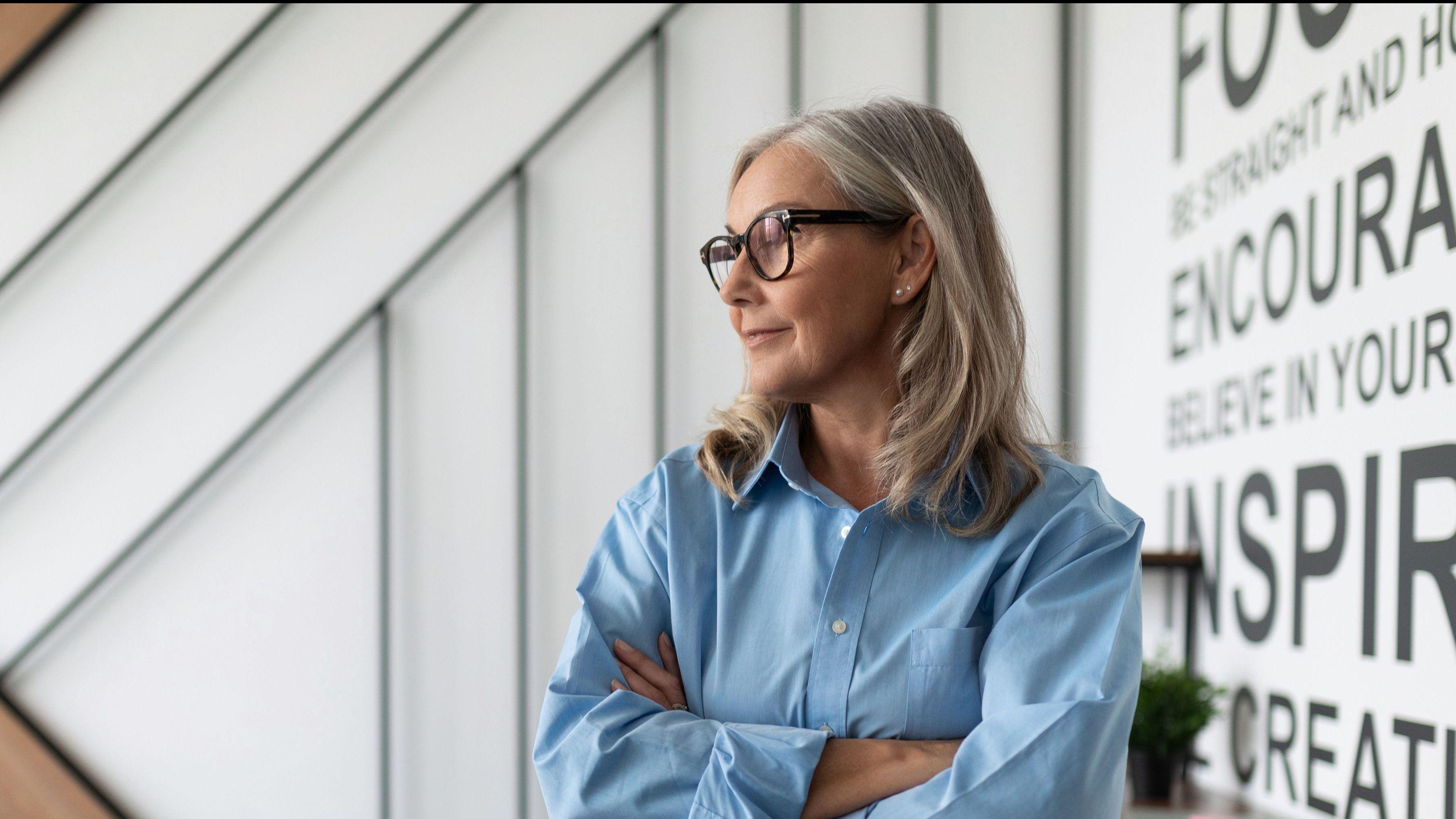 Frau mit langen grauen Haaren und Brille steht mit verschränkten Armen in einem modernen Büro vor einer Wand mit motivierenden Schriftzügen.